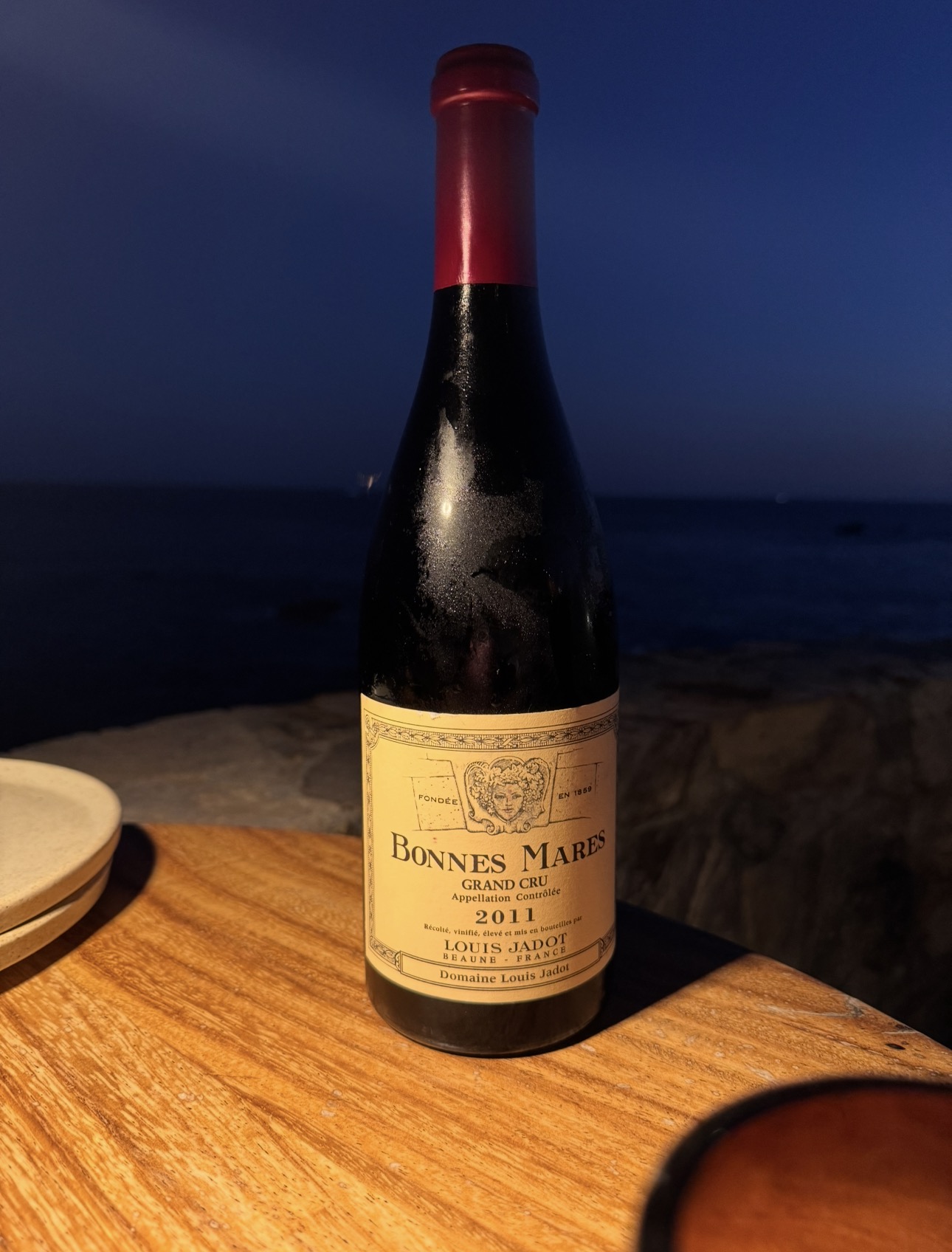 Louis Jadot Bonnes Mares Grand Cru 2011 bottle on a warmly lit wooden table against a dark blue background